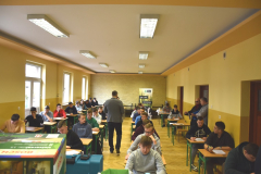 Młodzież siedzi w ławkach przy których stoi mężczyzna