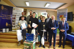 Mężczyźni, kobiety oraz uczniowie z pucharami w rękach pozują na stojąco do zdjęcia, w tle banery