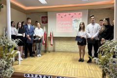 Uczniowie stoją na scenie, obok stoją kwiaty w wazonach, w tle godło, flagi