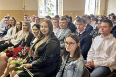 Uczniowie siedzą na krzesłach, na kolanach trzymają kwiaty