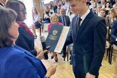 Nauczycielka wręcza uczniowi świadectwo, w tle siedzi grupa osób