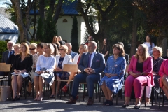 Nauczyciele siedzą, w tle drzewa, budynek.