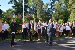 Poczet sztandarowy szkoły oraz uczniowie stoją, w tle drzewa, kwiaty