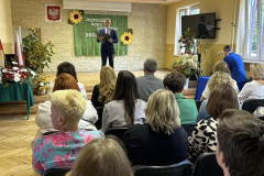 Mężczyzna stoi na scenie przy mikrofonie, pozostali uczestnicy apelu siedzą na krzesłach, w tle napis rozpoczęcie roku