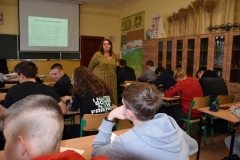 Młodzież siedzi w ławkach, kobieta stoi, w tle szafy