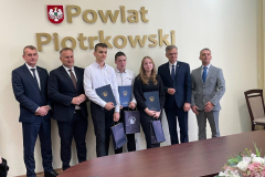 Uczniowie i mężczyźni pozują na stojąco do zdjęcia, nad nimi herb i napis Powiat Piotrkowski