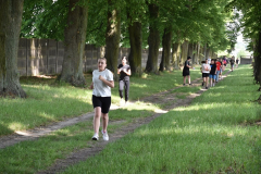 Dziewczyna biegnie ścieżką wśród drzew, w tle inne osoby