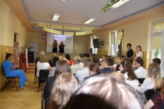 Na scenie stoją dwie uczennice, z boku stoi poczet sztandarowy, pozostali uczestnicy akademii siedzą, w tle godło, ekran rzutnika