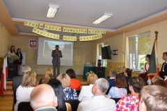 Wicestarosta Powiatu Piotrkowskiego stoi na scenie i mówi, pozostali uczestnicy akademii siedzą, w tle godło, ekran rzutnika, okno