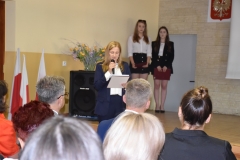 Zastępca wójta Gminy Czarnocin stoi z mikrofonem w ręku, w tle kwiaty, głośnik, dwie uczennice stoją