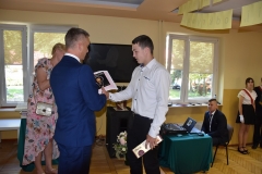 Dyrektor szkoły wręcza uczniowi nagrodę i świadectwo, w tle okna