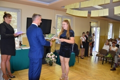 Dyrektor szkoły wręcza uczennicy książkę, w tle okno, telewizor, obok stoi wychowawczyni z książka