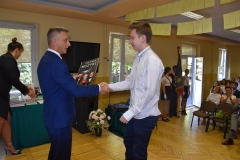 Dyrektor szkoły wręcza uczniowi książkę, w tle okno, telewizor, kwiaty w wazonie