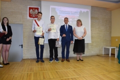 Dyrektor, nauczycielka i uczniowie stoją z dyplomami w ręku, w tle godło, zdjęcie budynku szkoły