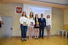 Dyrektor, nauczycielka i uczennice stoją z książkami w ręku, w tle godło oraz zdjęcie budynku szkoły