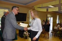 Wicestarosta Powiatu Piotrkowskiego wręcza uczennicy certyfikat ukończenia szkolenia, w tle telewizor, okno, bukiet kwiatów w wazonie