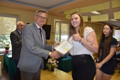 Wicestarosta Powiatu Piotrkowskiego wręcza uczennicy certyfikat ukończenia szkolenia, w tle telewizor, okno, bukiet kwiatów w wazonie