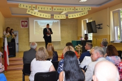 Przewodniczący Samorządu Szkolnego stoi, nauczyciele siedzą, w tle zdjęcie budynku szkoły, godło