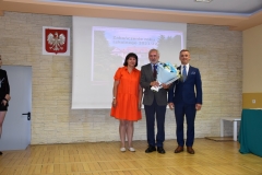 Dyrektor szkoły, wicedyrektor szkoły oraz kierownik szkolenia praktycznego z kwiatami w ręku stoją, w tle godło, zdjęcie budynku szkoły