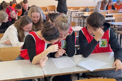 Młodzież siedzi pochylona nad kartkami leżącymi na ławkach