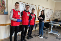 Uczniowie w czerwonych koszulkach stoją w tle plakaty informacyjne