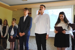 Uczniowie ubrani w strój galowy stoją, w tle ekran rzutnika i godło