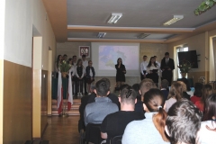 Uczniowie ubrani w strój galowy stoją na scenie, widownia, w tle ekran rzutnika i godło