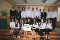 Uczniowie ubrani w strój galowy stoją, uczennice siedzą na schodach , z boku stoi w koszu bukiet biało-czerwonych kwiatów