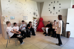 Chłopcy siedzą na krzesłach przed nimi stoi kobieta, w tle napisy, zegar