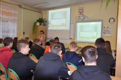 Uczniowie siedzą na krzesłach, nauczyciel stoi, w tle napis znamię znam je