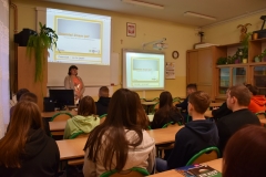 Uczniowie siedzą w ławkach, nauczycielka stoi, w tle szafy