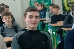 Uczeń w czarno-szarej bluzie siedzi, w tle inni uczniowie