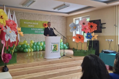 Mężczyzna stoi przy mónicy, z boku stoją kwiaty z tyłu na ścianie napis Rozpoczęcie roku szkolnego