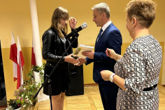 Dyrektor szkoły i wychowawczyni wręczają książkę uczennicy, w tle białoczerwone flagi