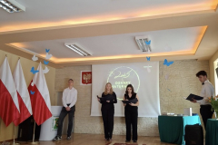 Uczniowie stoją na scenie, obok są flagi, przed nimi rozłożony chodnik