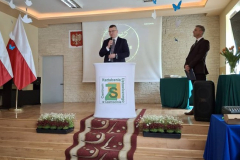 Mężczyzna stoi przy mównicy, obok stoi dyrektor szkoły w tle flagi biało-czerwone