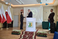 Mężczyzna gratuluje kobiecie, obok stoi drugi mężczyzna, w tle flagi biało-czerwone