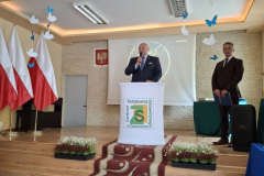 Mężczyzna z mikrofonem w ręku stoi przy mównicy, obok stoi drugi mężczyzna, w tle biało-czerwone flagi