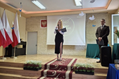 Kobieta z mikrofonem w ręku stoi na scenie, obok stoi mężczyzna, w tle białoczerwone flagi