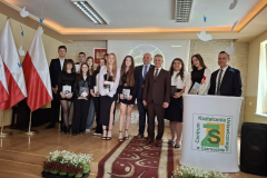 Absolwenci pozują do zdjęcia z nauczycielami, obok stoją białoczerwone flagi i mównica