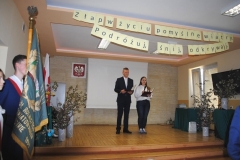 Uczniowie stoją na scenie w tle godło szkoły, ekran rzutnika