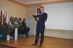 Dyrektor szkoły przemawia stojąc, w tle bukiety kwiatów i ściana, na ścianie godło