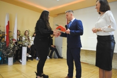 Dyrektor szkoły i wychowawczyni wręczają nagrodę i świadectwo uczennicy, w tle ściana