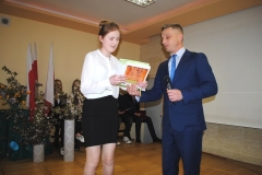 Dyrektor szkoły i wychowawczyni wręczają nagrodę i świadectwo uczennicy, w tle ściana