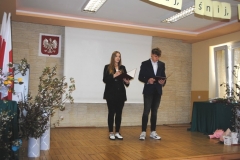 Uczniowie stoją i przemawiają, w tle ściana, bukiety kwiatów, godło