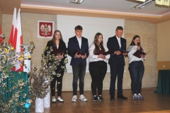 Uczniowie stoją i przemawiają, w tle ściana, bukiety kwiatów, godło